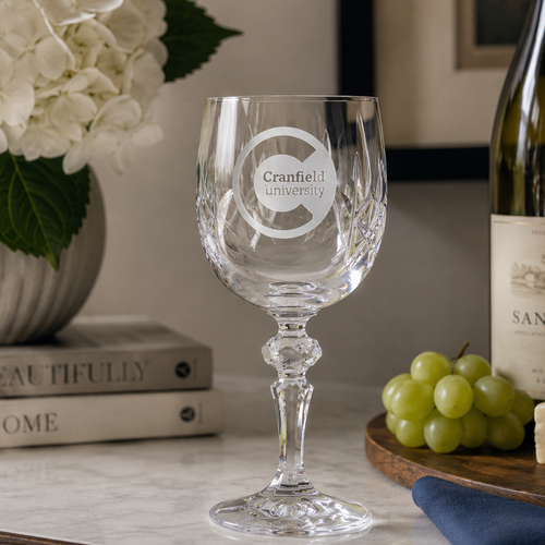 Clear wine glass with Cranfield University logo on a table with grapes and a bottle of wine.