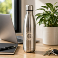 Silver water bottle on a desk with a laptop, notebook, pen, keys, and plant in the background.